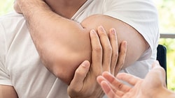 Man holding his elbow in pain during a consultation, possibly recovering from a bicep tendon rupture or repair.