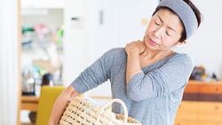 A woman in a gray shirt, holding a laundry basket and rubbing her neck, stands among wooden furniture.