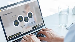Person using a laptop showing a medbridge dashboard with circular graphics, next to a water glass and lamp on a desk.