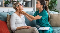A healthcare worker in scrubs uses digital tools to check an older woman's neck on a couch at home, using medbridge.