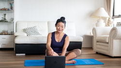 A woman smiles as she sits cross-legged on a yoga mat in her living room, looking at a medbridge course on her laptop.