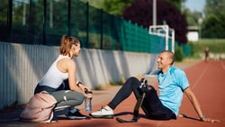 A male runner with a prosthetic blade leg sits on a track, smiling at a female coach.