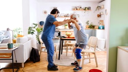 Male OT helping an elderly man practice a sit-to-stand transition in a home kitchen setting.