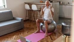 Man sitting on an exercise ball, drinking water, with a yoga mat and laptop nearby in a living room.