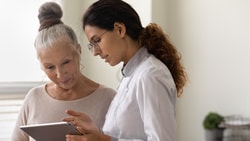 A woman in a lab coat shows a tablet to an older woman in a well-lit room, demonstrating medbridge.