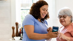 Two women in a kitchen, one sharing her smartphone screen with the other amidst cooking utensils and cutting boards; medbridge styling.