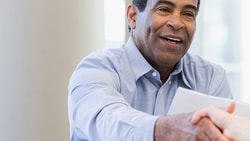 Two people are shaking hands across a table while another person sits nearby, smiling.