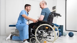 A therapist kneeling and assisting a wheelchair user with positioning in a bright, modern therapy clinic.