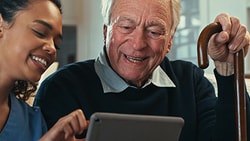 A young woman shares a tablet with an elderly man holding a cane, both smiling in a living room.
