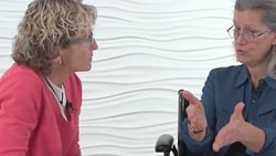 Two people conversing, one gesturing, against a textured white wall backdrop.