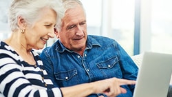 An older couple uses a laptop together in a bright room, with the woman pointing at the screen.