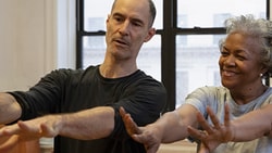 Two people exercise indoors with arms extended, behind them are a window and exercise balls.