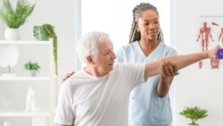 A caregiver helps an elderly man with a shoulder exercise in a bright room with anatomical charts and plants.
