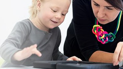 A child and an adult with a colorful necklace focus on a black circular object on a table during Medbridge's activity.