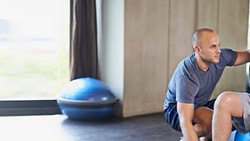 An older man is assisted by a younger man while sitting on an exercise ball in a gym, as seen on medbridge.