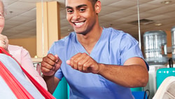 An elderly person exercises with a red resistance band, assisted by a smiling healthcare worker in medbridge scrubs.