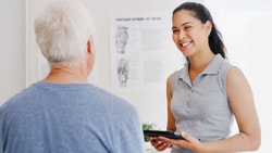 A woman with a tablet smiles at an older man in a medical office, showing the benefits of medbridge digital healthcare.
