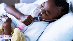 A child in bed with a nebulizer mask and holding a teddy bear attached to an IV drip.