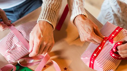 Two people in sweaters wrap gifts with red and white striped paper at a table.