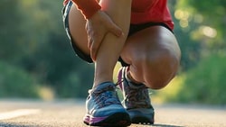A person kneeling on a road holds their leg in discomfort against a natural background.