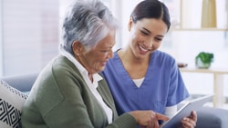 A healthcare professional in blue scrubs smiling while showing a digital tablet to an older woman in a home setting.