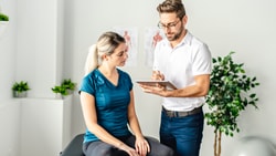 A woman sits on an exam table as a male healthcare professional takes notes, illustrating patient care using medbridge software.