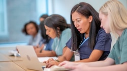 Healthcare professionals in blue and green scrubs are seated at a table, focused on learning. Two women in the foreground are using a laptop and a book. The setting is a brightly lit room. The atmosphere is professional and focused.