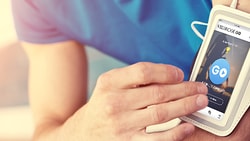 A person in athletic wear uses a smartphone in a white armband holder with the medbridge music app displaying a blue GO button.