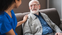 A caregiver, appearing to be a younger woman with dark hair and wearing a blue scrub top, gently places her hand on the shoulder of an older adult man with white hair and a beard, who is seated in a brown armchair and smiling. The setting appears to be indoors, possibly a care facility or the man's home. The interaction suggests a caring and supportive relationship.