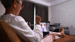 An older woman with short grey hair and red-rimmed glasses sits comfortably on a brown bed, looking at a laptop resting on her lap. She is wearing a white long-sleeved shirt and light pink pants. On the laptop screen, a younger woman in a light purple shirt smiles and holds up a closed fist in a gesture of encouragement or success. The background shows a bedroom setting with dark curtains, white walls, and a bedside table. The scene suggests a virtual connection or online interaction between the two women.