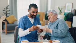 A caregiver in blue scrubs assists an elderly woman with reading at a table, featuring cups and a notepad.