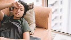 Someone resting on a sofa with a blanket by a window, hand on forehead.