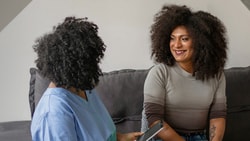 Two curly-haired individuals converse on a couch; one has a clipboard, and the other is smiling.