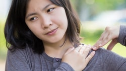 Individual in a long-sleeve shirt outdoors, touching their shoulder with a thoughtful look.