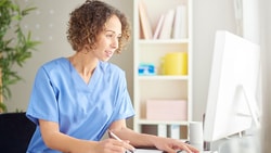 A person in blue scrubs writes at a desk with a computer, plant, and shelves visible.