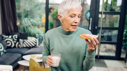 A woman with short gray hair holds a grapefruit slice and a glass of water in a living room with large windows.