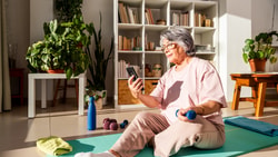 Older woman on a yoga mat holds a dumbbell and checks her phone in a bright room with medbridge training gear and plants.
