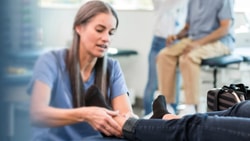 A physical therapist assesses a patient's ankle in a clinic; medbridge session visible in background.