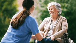 A caregiver in blue scrubs smiles beside an elderly woman in a wheelchair outdoors.