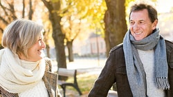 A man and woman in scarves and coats smile at each other while walking on a path lined with autumn leaves.