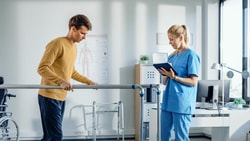 A man in a mustard shirt uses parallel bars as a woman in blue scrubs takes notes at a Medbridge clinic.