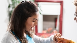 A young woman assists an older woman with a bracelet indoors, smiling near a mirror and plant.