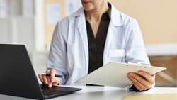 Clinician holds a clipboard while entering patient outcome data into a laptop at a clinic workstation