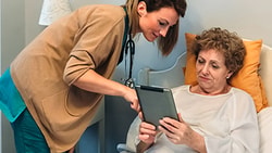 A healthcare professional presents information on a tablet to an elderly patient in bed, exemplifying medbridge's digital care.