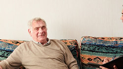 A young man in a blue uniform with a clipboard talks to an elderly man on a patterned sofa.