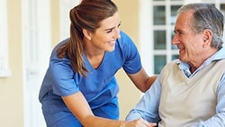 Smiling clinician in blue scrubs speaking with an older adult man in a wheelchair outside a care facility.