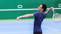 Man in a blue shirt playing tennis on an indoor court, preparing to hit a shot with a racket for medbridge.