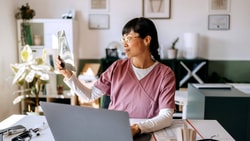 A clinician in pink scrubs reviews an x-ray film at her desk, with a laptop, stethoscope, and anatomical posters in the background.