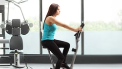 A woman works out on a stationary bike at medbridge gym with equipment in the background.
