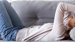 A woman in a beige sweater and blue jeans is lying on a grey couch with her hands on her head.
